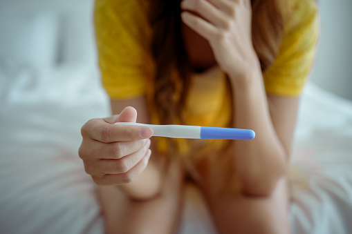 Young asian young woman feeling shock after look at positive Pregnancy test. Disappointed unplanned pregnancy girl sit on bed with upset and sad face for problem don't want to pregnant.