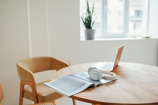 Laptop, headphones and magazine on a round wooden table next to a comfortable beige armchair. A green potted houseplant on the windowsill. A cozy freelance workplace in a minimalistic interior.