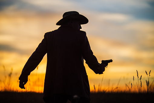 Silhouette of a cowboy in a hat with a revolver against a dramatic sunset sky. Western concept. Life in the wild west.