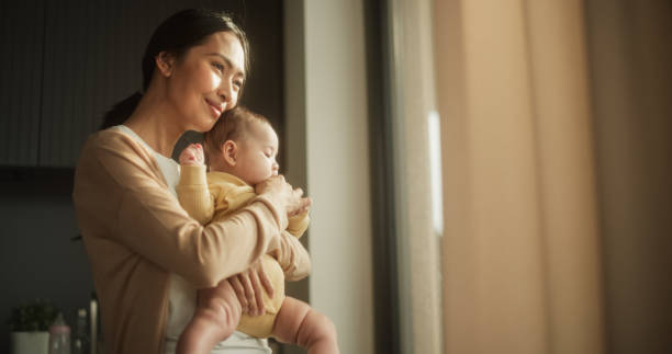 家の窓のそばに立っている間、赤ちゃんを腕に抱く美しいアジアの若い母親。幸せな女性、母性に不慣れ、子供を抱きしめ、一緒に絆の瞬間を楽しむ - male nurse 写真 ストックフォトと画像