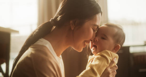 concetto di affetto materno: ritratto ravvicinato di un'affettuosa madre asiatica che tocca il naso con il suo bambino in un'accogliente camera da letto illuminata naturalmente. atmosfera calda che riflette il momento commovente - mother new baby nursery foto e immagini stock