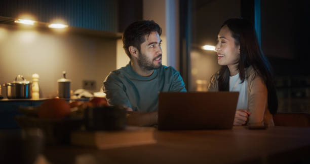 istock Portrait of Young Couple Using Laptop Computer While Sitting in the Kitchen at Home And Talking. Engaged Couple Doing Online Shopping for their Wedding Preparations, Discussing Different Options. 1514021877