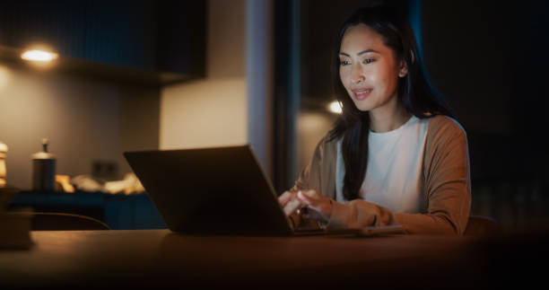 istock Beautiful Authentic Asian Woman Sitting at a Table in a Cozy Kitchen and Using Laptop Computer at Home at Night. Female Smiling and Browsing the Internet, Checking Videos on Social Networks. 1514021893