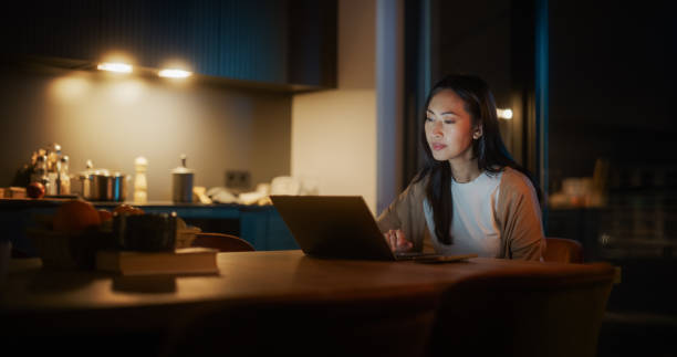 istock Beautiful Authentic Asian Woman Sitting at a Table in Cozy Kitchen and Using Laptop Computer at Home at Night. Female Smiling and Chatting on the Internet and Social Media, Doing Scientific Research. 1514022418