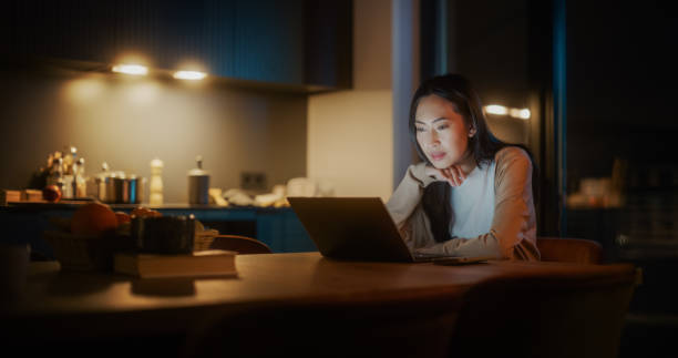 istock Beautiful Authentic Asian Woman Sitting at a Table in a Cozy Kitchen and Using Laptop Computer at Home at Night. Female Smiling and Chatting on the Internet and Social Media, Doing Research 1514024033