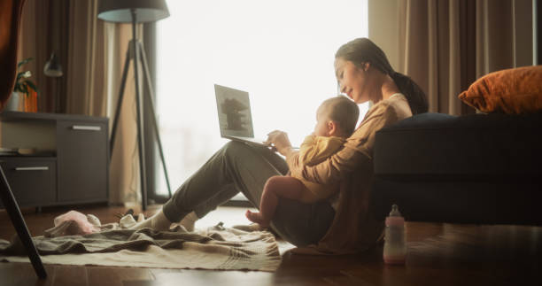 Working from Home: Portrait of Young Asian Mother, Holding her Cute Baby and Working on Laptop Computer During Day. Successful Female Architecte Balancing Life and Work while Doing Job Remotely. trabajar desde casa: retrato de una joven madre asiática, sosteniendo a su lindo bebé y trabajando en una computadora portátil durante el día. arquitecta femenina exitosa que equilibra la vida y el trabajo mientras realiza el trabajo de forma remota.