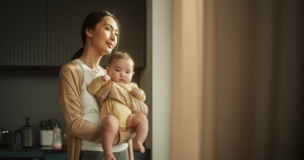 아름 다운 젊은 아시아 어머니는 집에서 창문 옆에 서있는 동안 그녀의 팔에 아기를 안고 있습니다. 행복한 여자, 모성에 처음, 웃고 그녀의 아이와 함께 햇빛의 따뜻함을 즐기는 - human pregnancy baby shower image color image 뉴스 사진 이미지