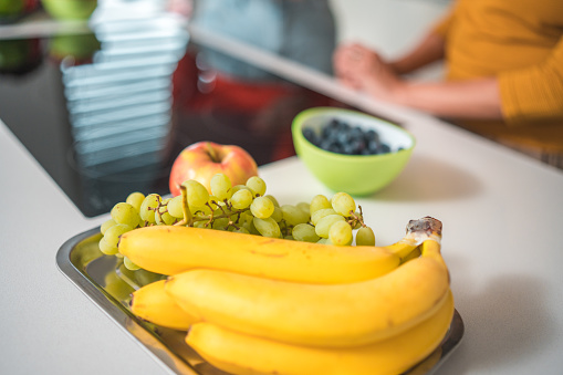 Close up of a pile of fresh bananas, grapes and an apple, all on a tray in a modern kitchen. A woman in blurred background.