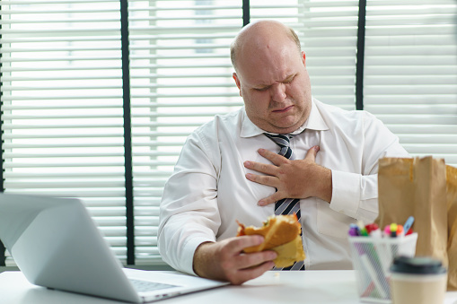 Unhealthy oversize fat man employee working in the office overtime and always eating junk food, obese businessman enjoy eating cheeseburger while working in office.