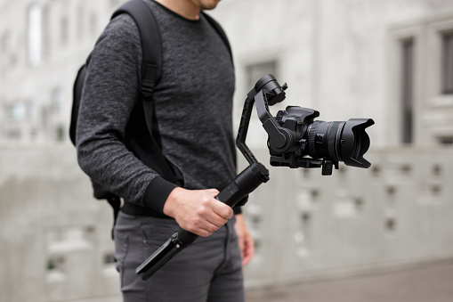 videography, filmmaking and creativity concept - close up of modern dslr camera on 3-axis gimbal stabilizer in male hands over concrete building background