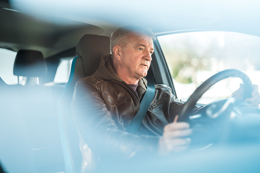 Through window shot of a man with gray hay. He is seated inside his electric car, holding the steering wheel.