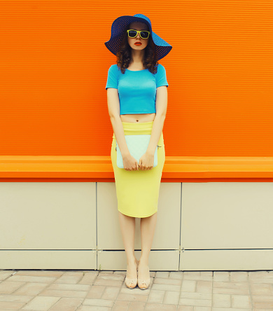 Beautiful young woman in full-length posing wearing summer straw hat, skirt and handbag clutch on city street