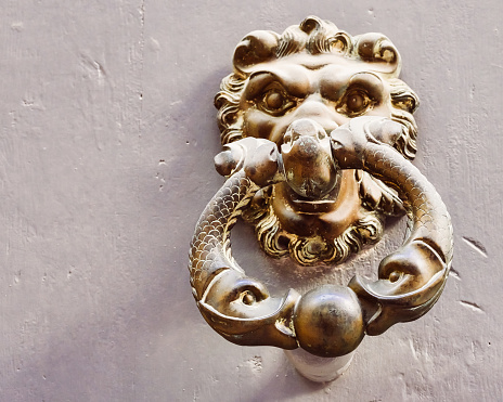 Old weathered brass il-Habbata door knocker featuring an oriental dog head and figures hanging from its mouth, set on a brown wooden door in Malta.