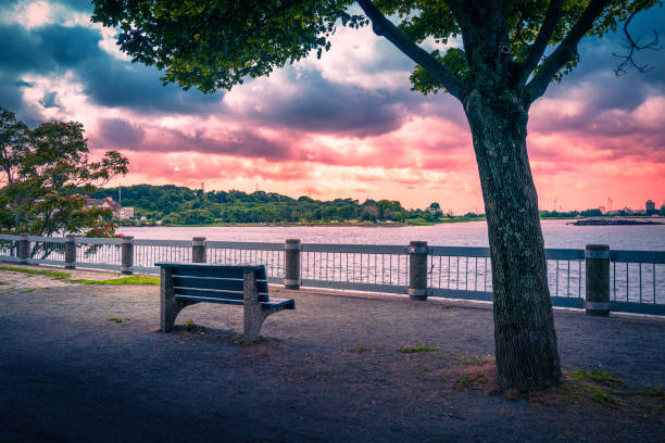 Tranquil sunset in the fenced river park with an empty rustic wooden bench and slanted old tree. Indian Point Park along Seekonk River in Providence, Rhode Island.