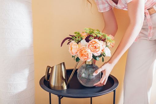 Woman puts vase with bouquet of flowers on table at home. Floral arrangement with orange roses. Interior and summer decor