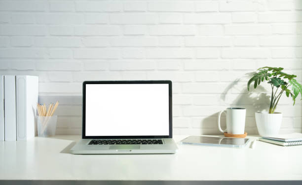 istock Laptop computer, coffee cup and book on white table against brick wall. Blank screen for your advertise text. 1514219340