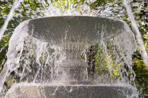 Close-up granite bowl in fountain in splashes and jets of water on greenery of park background, comfortable urban environment. large round stone fountain made of granite