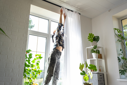 Woman hangs transparent tulle curtains on large windows in the house inside the interior. Spring cleaning, tidying up.