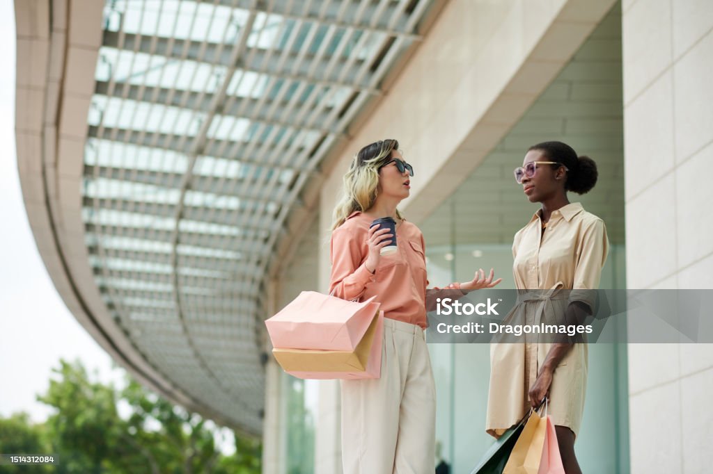 Stylish Women Standing at Mall Entrance Stylish young women standing at shopping mall entrance and discussing news 20-24 Years Stock Photo
