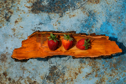 Three strawberries on the red plate of oak bark. Blue rusty metal background. Copy space.