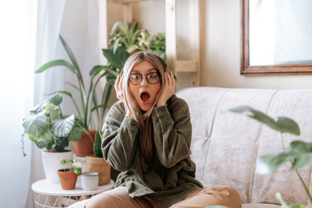 mulher chocada com a boca aberta nos óculos ouvindo música em fones de ouvido e relaxando em casa. menina frustrada sentada no sofá na sala de estar com plantas. conceito de selva urbana. - business mobile phone telephone women - fotografias e filmes do acervo