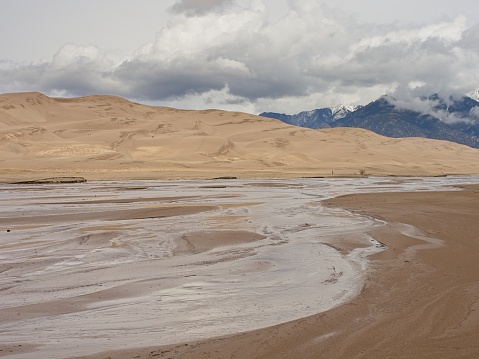 The meandering Buck creek in front of the huge sand dunes in Great Sand Dunes national park. Moody skies and the Sangre de Cristo mountains rise in the distance.