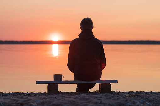 Man is sitting on a wooden bench on the shore of the sunset sea. The background is blurred.