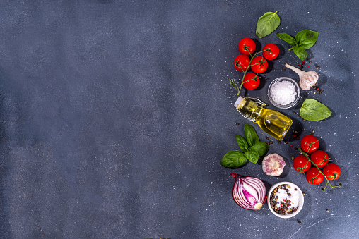 Black cooking background with useful cooking italian Mediterranean ingredients - tomatoes, basil leaves, greens, olive oil, salt, pepper, garlic, flat lay black concrete table top view copy space