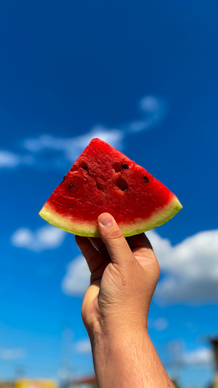 A hand holding a slice of watermelon, with a blue sky from behind