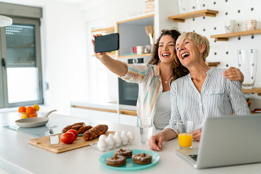 Mother and daughter laughing while using mobile phone at kitchen counter.