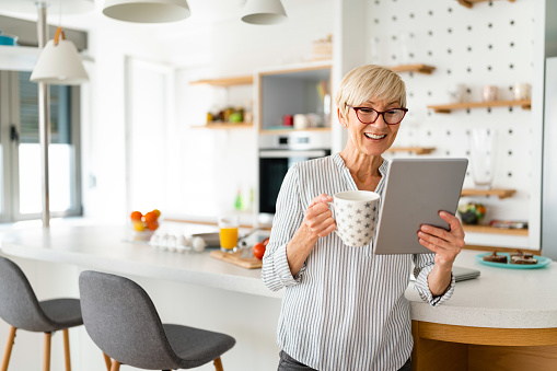 Smiling mature woman drinking coffee while reading online news on digital tablet and leaning on table in the morning.