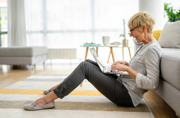 istock Satisfied excited elderly woman casually dressed, typing on laptop while sitting on the floor in living room 1514695118