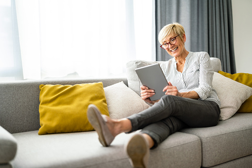 Widely smiling cheerful senior woman reading news on digital tablet while sitting on couch in living room.