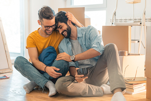 Cute homosexual couple sitting on floor and enjoying new home while hugging and drinking coffee.