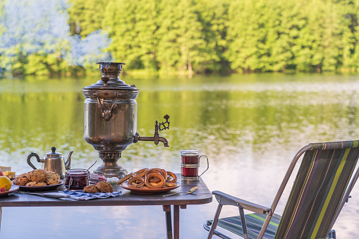 Vintage metal tea samovar with white smoke and food on the table near the calm water lake in green forest at morning, Ukraine. Nature food concept