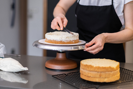 Skilled female confectioner smearing organic cake layers with knife standing at table in modern kitchen at home housewife enjoying cooking delicious confectionery for customers