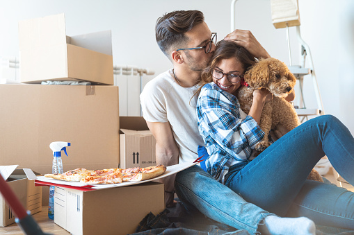 Young couple with a dog happy in their new family home sitting on the floor having pizza surrounded with cardboard boxes.