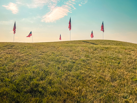 Six American Flags Flying High Perched at the Top of a Hill. Green hill in the forefront of view, pale blue morning skies with clouds in the background.