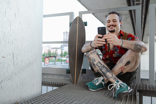 A Latin American man in his 30s is surfing the net on his mobile phone while spending time in the city. His skateboard is next to him.