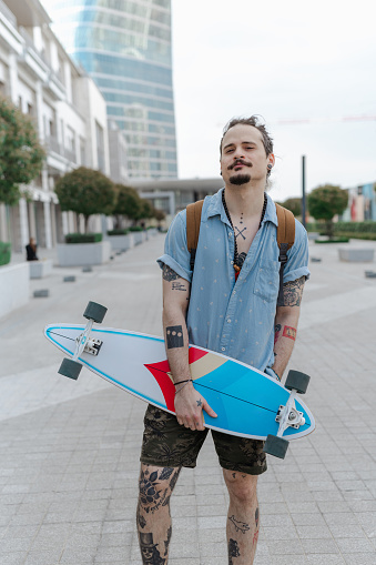 Portrait of an urban Latin American man with a tattooed body holding his skateboard and looking at the camera