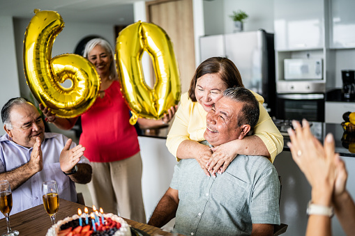 Senior man celebrating his birthday with friends while wife is embracing him at home
