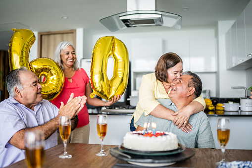 Senior man celebrating his birthday with friends while wife is embracing him at home