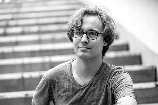 Black and white portrait of  26 years old man with eyeglasses sitting on stairs, background with copy space, full frame horizontal composition