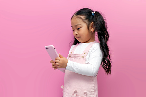 little asian girl in pink sundress uses smartphone and chooses online on pink isolated background, korean preschool child holds phone and clicks on the screen