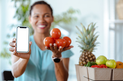 African American woman holding a cardboard box full of organic food and vegetables and showing blank smart phones screen