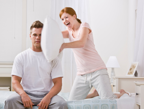 A cute and young redhead hitting her boyfriend with a pillow.  She has a joyful look on her face.  His expression is serious.  Horizontally framed shot.