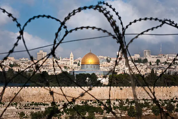 The Old City of Jerusalem, including the Dome of the Rock and various church steeples, seen through coils of razor wire, illustrating the Holy Land's history of division and conflict.