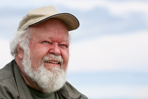 A closeup of a senior male with white beard and hair, in a baseball cap, isolated against sky.