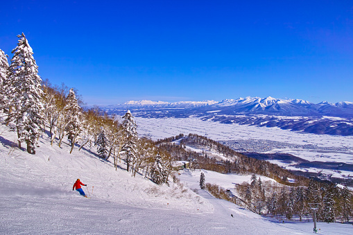 Furano, Hokkaido, Japan - January 30, 2023: Daisetsuzan and the Tokachidake mountain range seen from the Furano ski resort on a clear day, skiers skiing on the high slopes of the intermediate course from the highest point in the Furano zone
