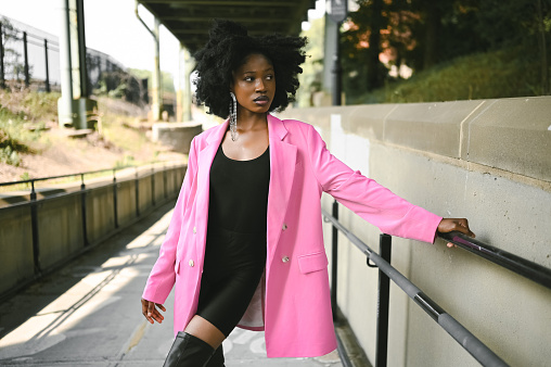 Fashion outdoor street style portrait of Beautiful young African American woman posing outside on urban city landscape summer day wearing pink jacket. Attractive black female, diversity concept